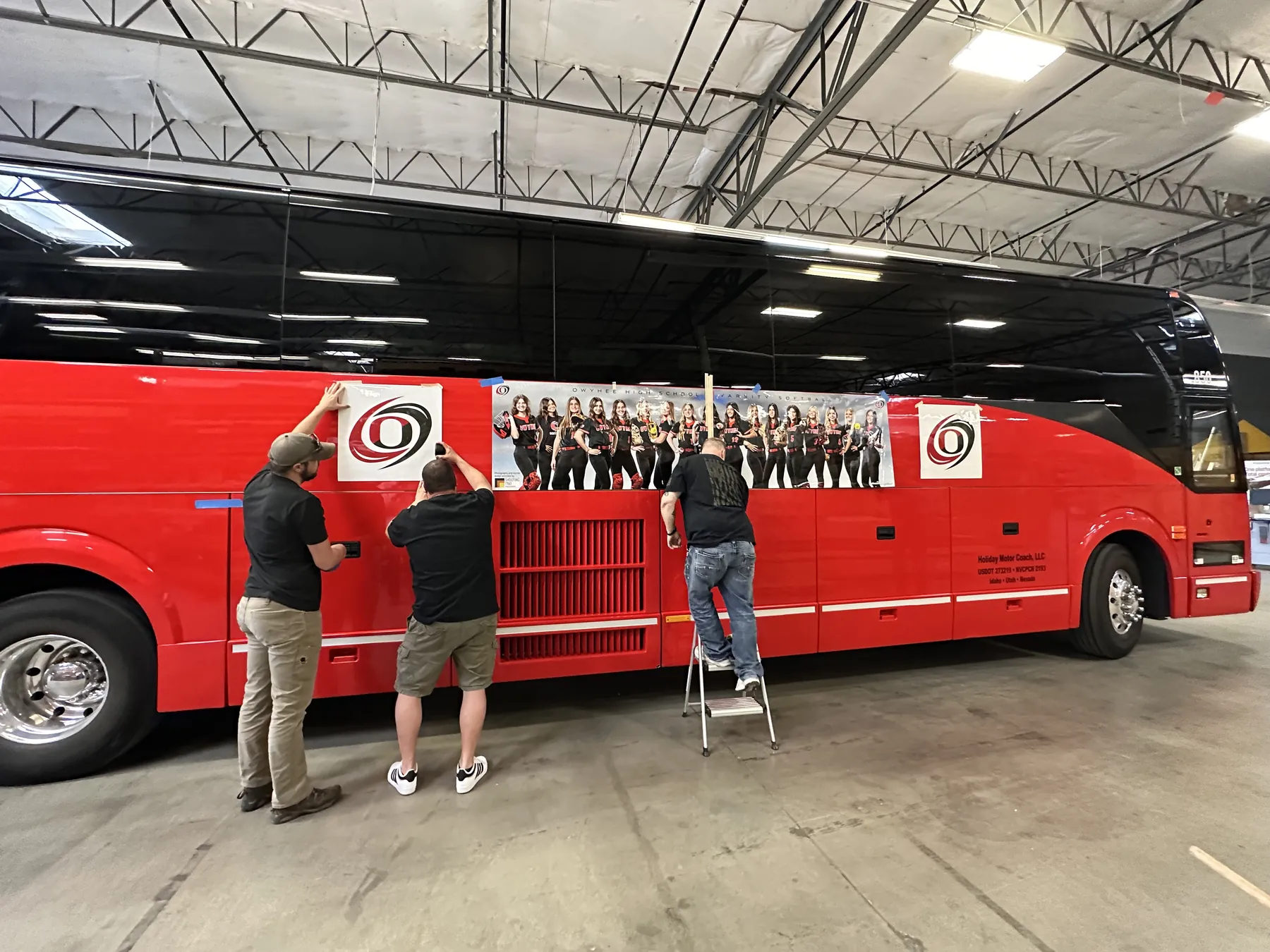 Owyhee High School varsity softball team banner being applied as a wrap on the side of a red charter bus before the state tournament