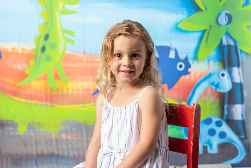 Preschool girl portrait in front of colorful dinosaur backdrop at photo day - school photography Boise Meridian