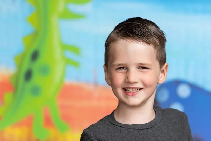 Smiling preschool boy headshot at photo day with hand-painted dinosaur backdrop - school photographer Meridian